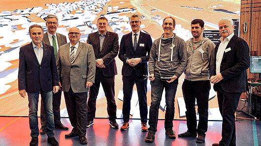 Gruppenbild vor einem LED-Wand mit einem Luftbild einer Kleinstadt und und der sie umgebenden Landschaft