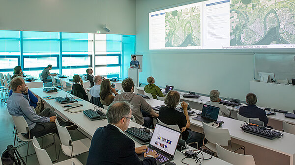 Photograph of HLRS auditorium made during the conference. A man stands at the podium with an aerial image of vineyards along a bend in the Neckar River.
