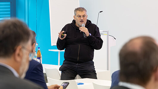 A man sits on a desk, holding a microphone and giving a speech at the front of an auditorium.