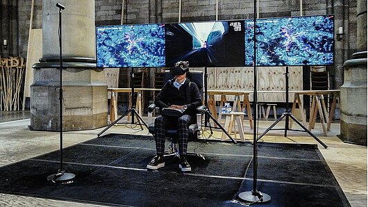 Person sits in chair wearing 3D headset, while screens show what he sees.