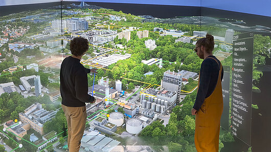 Two scientists in a CAVE visualization facility study a digital twin of the university campus, depicted in virtual reality from an aerial perspective.