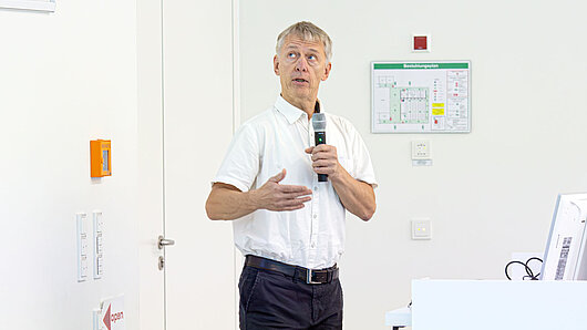A man holds a microphone while giving a speech, glancing at a screen on the wall.