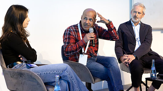 A panel discussion with three people. A man in the middle speaks into a microphone.