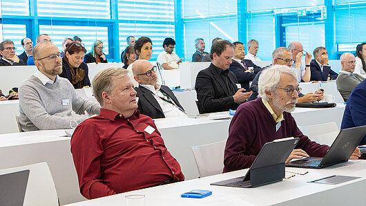 An audience sits at tables listening to a lecture in an auditorium.