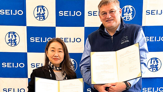 Photo of Prof. Myojo and Prof. Resch holding copies of the signed agreement in front of a wall with the Seijo University logo..