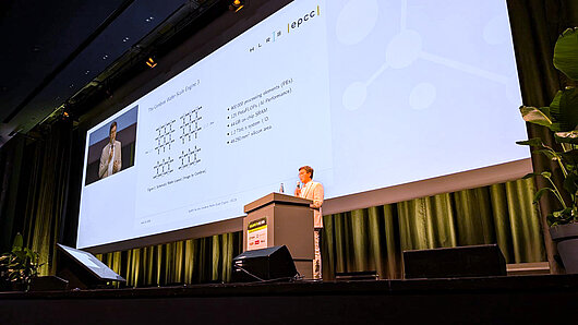 A man gives a lecture at a podium on a stage, with a large screen behind him.