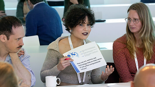 A woman holds up a sign during a group discussion that says "How can art, science, and technology help us make sense of and within computational culture?"