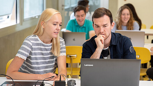 University students in a classroom work together on a laptop computer.