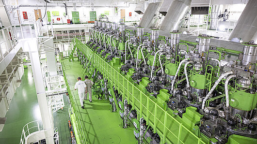 Photo of engine room with large machine and workers walking on a gangway.