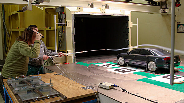 Photo of two scientists spraying a mist above a model car in a wind tunnel to see air flow. One wears goggles to observe the experiment in augmented reality.