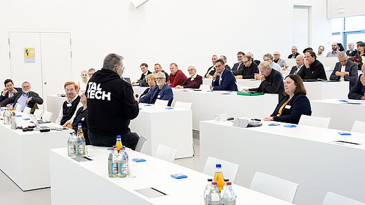 Photo of auditorium with audience. A speaker sits on a table at front of room.