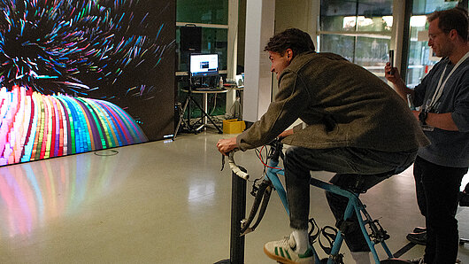 A man pedals a stationary bicycle, controlling a visualization on a large screen.