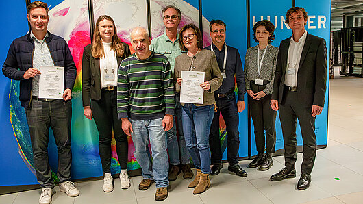 Mitglieder des HLRS-Teams und Auditoren halten Zertifikate vor dem Supercomputer Hunter in den Händen.