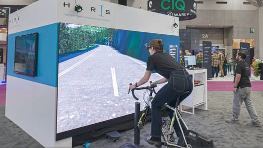 A woman wearing 3D glasses rides on a bicycle simulator, with a screen in front of her.