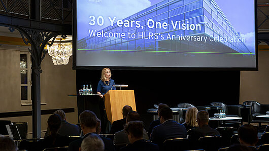 A woman speaks at a podium in front of an audience with a large screen in the background.