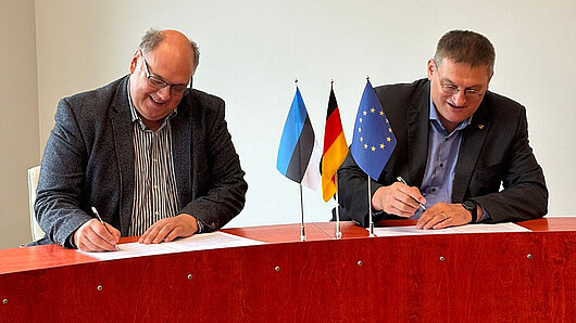 Erik Puura und Michael Resch sitzen an einem Tisch und unterzeichnen einen Vertrag. Auf dem Tisch liegen estnische, deutsche und EU-Flaggen.