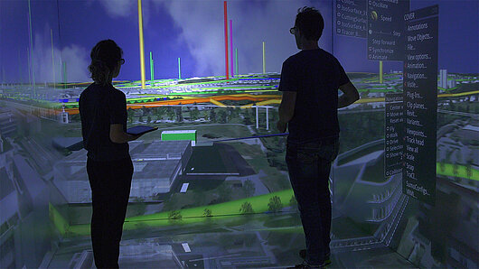 Two scientists observe a room-sized, projected visualization of the University of Stuttgart campus.